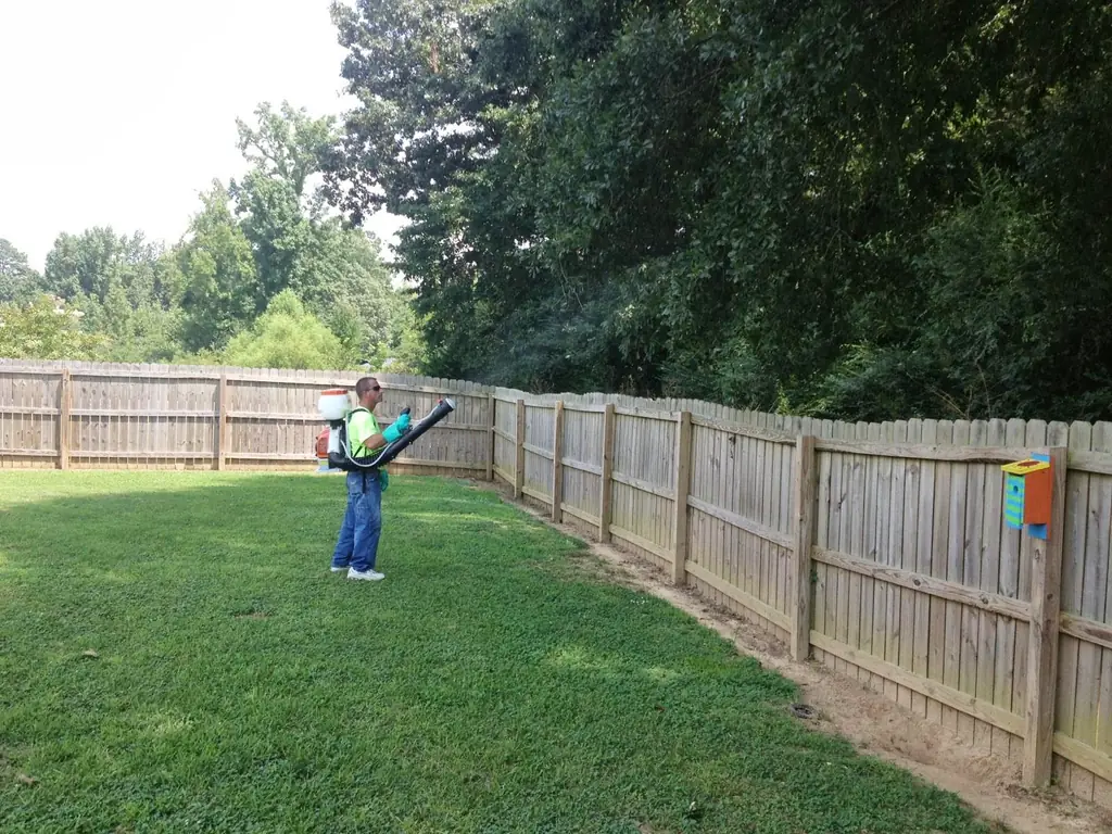 A Superior technician misting the barrier along a wooden fence line at the back of a Decatur yard, with dense treeline behind the fence